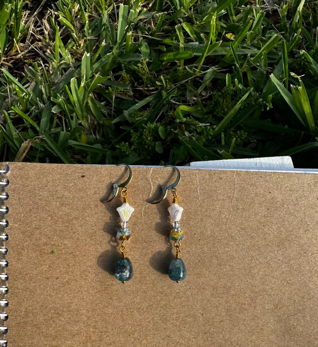 A pair of gold-colored three petal cathedral drop earrings with white and blue czech glass accents, displayed against a beige background.
