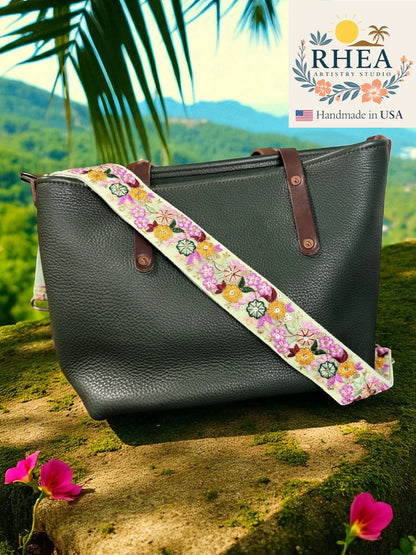 Black leather handbag with a floral strap against a scenic background, featuring the RHEA brand.