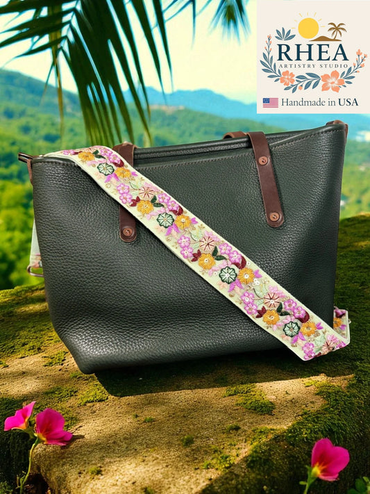 Black leather handbag with a floral strap against a scenic background, featuring the RHEA brand.