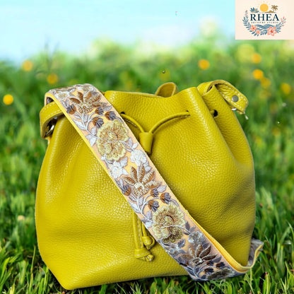 Light yellow handcrafted leather bucket bag with a wide  yellow floral embroidered strap, displayed on a tropical beach backdrop — Rose Bloom Collection by Rhea Artistry Studio.