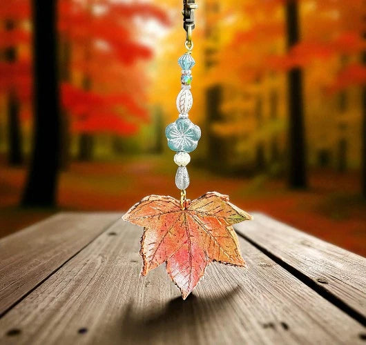 Decorative leaf charm with beads on a wooden surface with a blurred autumn forest background