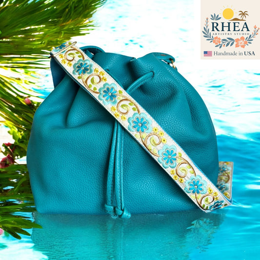 Blue leather bag with a decorative purse strap on water, RHEA Artisan Studio logo visible