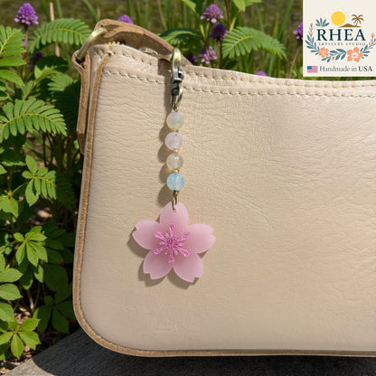 Beaded keychain with a pink flower charm attached to a beige bag, with greenery in the background. Floral beaded zipper charm with pink blossom pendant, handmade in USA
