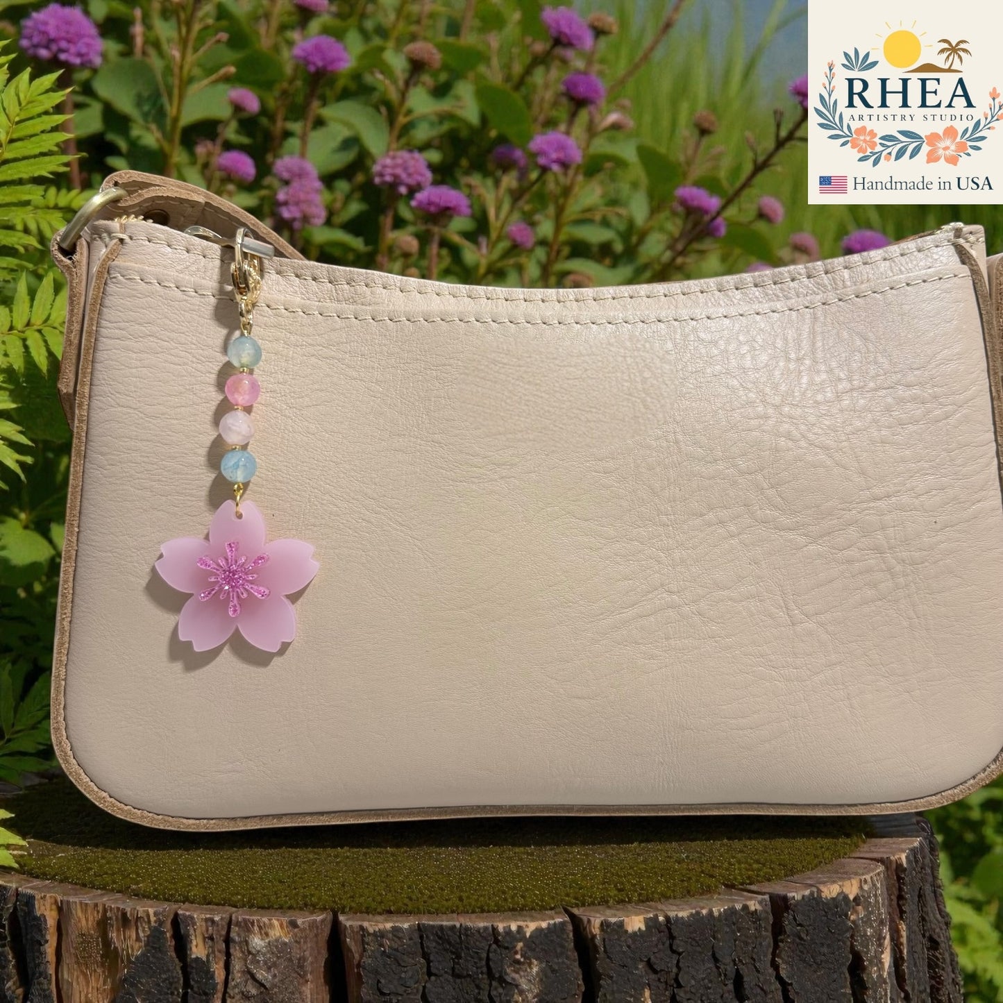 Beige handbag with a pink floral charm on a wooden stump with greenery in the background, featuring the RIHEA brand. Pastel pink cherry blossom bag charm with gold lobster clasp