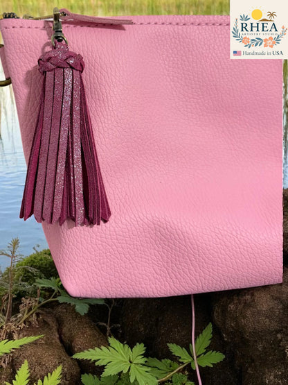 Pink leather clutch with a tassel on a natural background, featuring the RHEEA brand.