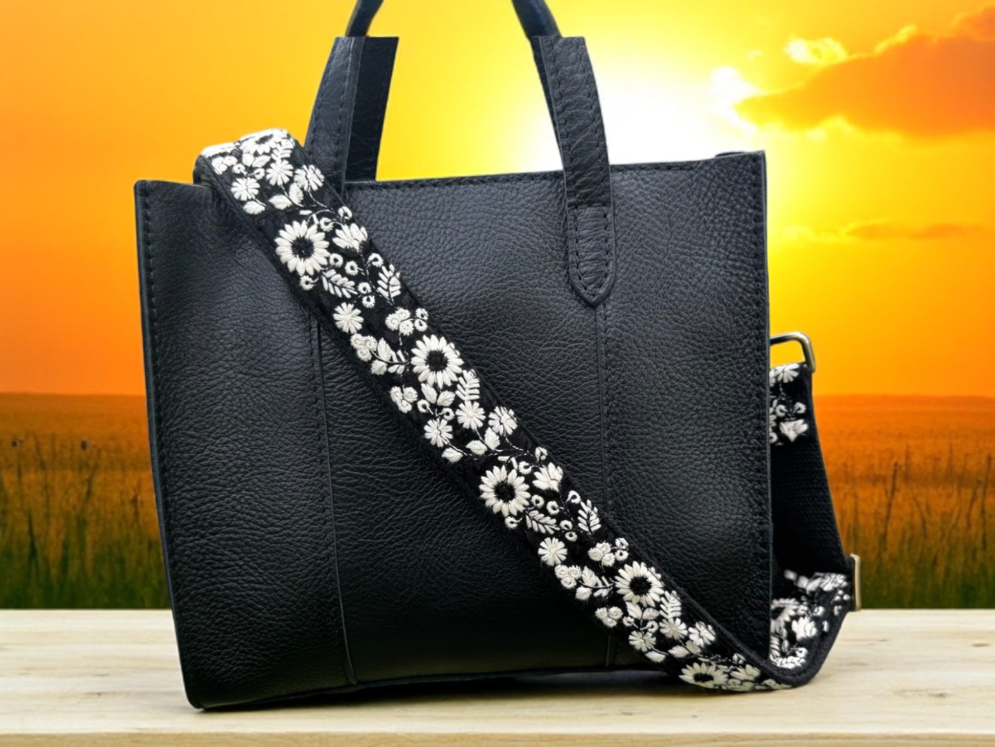 Elegant black fabric crossbody strap with detailed white wildflower embroidery, perfect for handbags and DSLR cameras. Ethically handcrafted with a modern flair against a sunset background