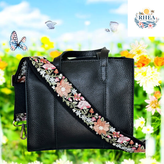 A structured black leather tote bag styled with a wide embroidered strap in black base fabric, adorned with detailed coral, blush, and white floral embroidery. The background bursts with springtime energy—meadow blooms, butterflies, and blue sky—enhancing the romantic, botanical charm of this artisan piec