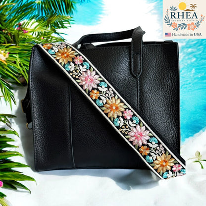 Black handbag with a floral purse strap on a beach-themed background, featuring the RHEA brand.