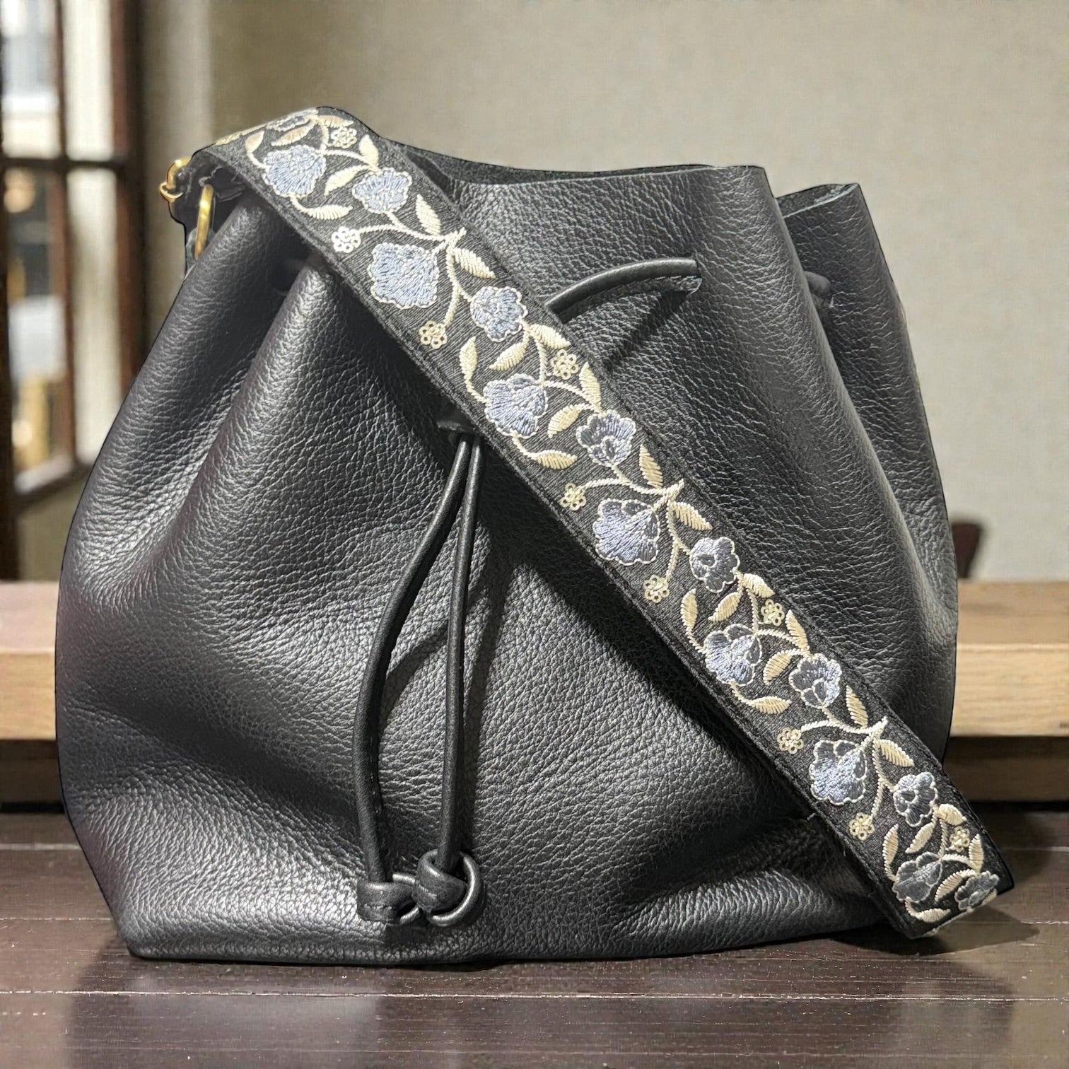 Floral embroidered bag strap measuring 1.5 inches wide, Black leather drawstring bag with floral embroidery on a wooden surface