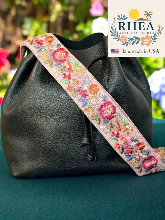 Black leather bag with a floral bag strap on a green surface, Rhea Artistry Studio logo visible.