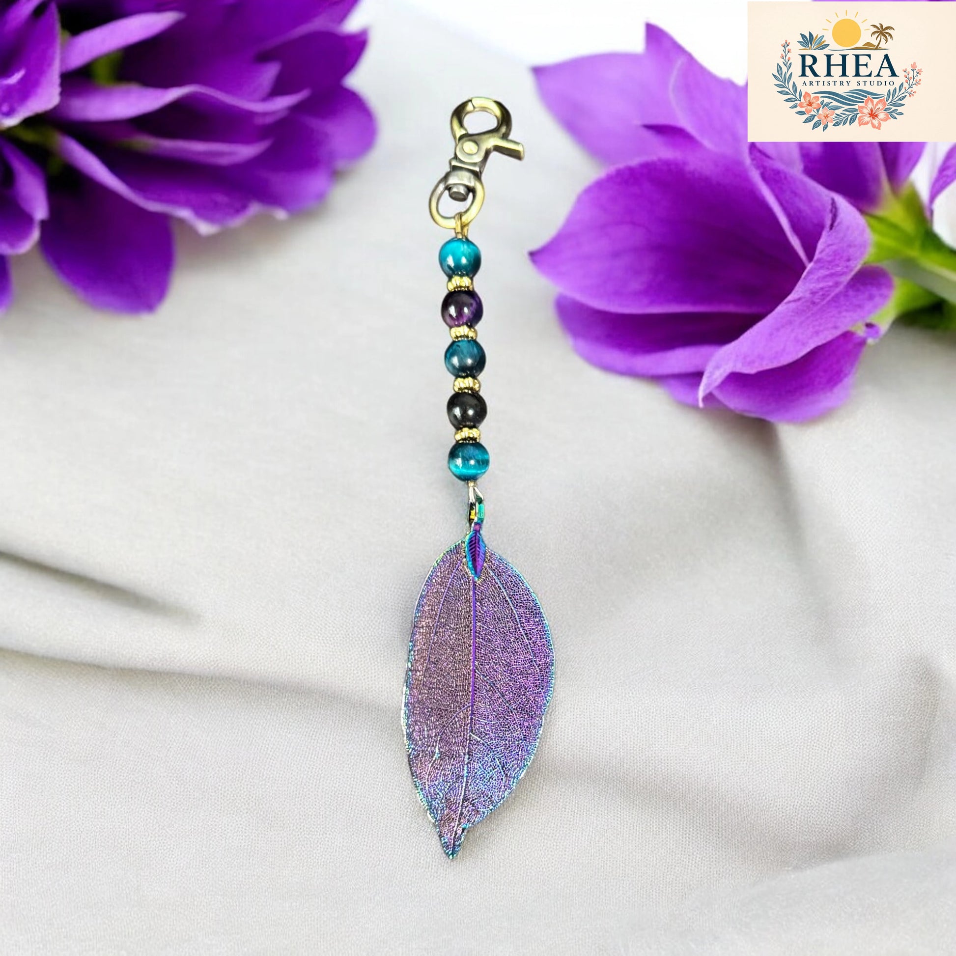 Purple leaf-shaped keychain with beads on a white fabric background with purple flowers.