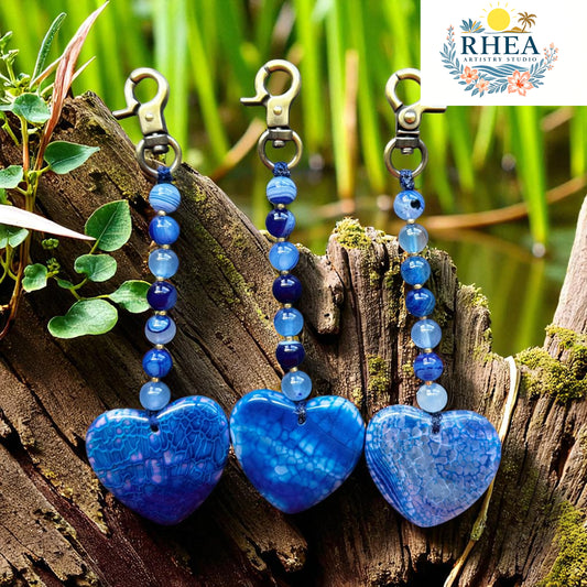 Blue heart-shaped keychains with beads on a wooden log with greenery in the background, featuring the RHEA brand.