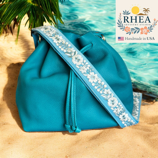 Blue handbag with floral purse strap on sand near water, RHEA Artistry Studio logo visible
