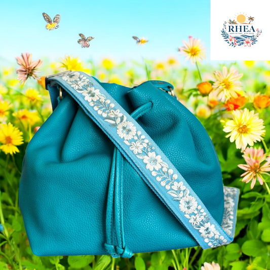 A teal leather bucket bag featuring the Bluebell Frost strap—handmade with powder blue fabric and white floral embroidery, shown on a meadow
