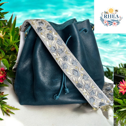 A 2-inch wide strap featuring soft blue floral embroidery with gold accents on a silver base, displayed on a dark teal leather bag