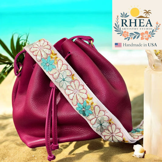 Pink leather bag with floral strap on a beach background, featuring RHEA Artistry Studio logo.
