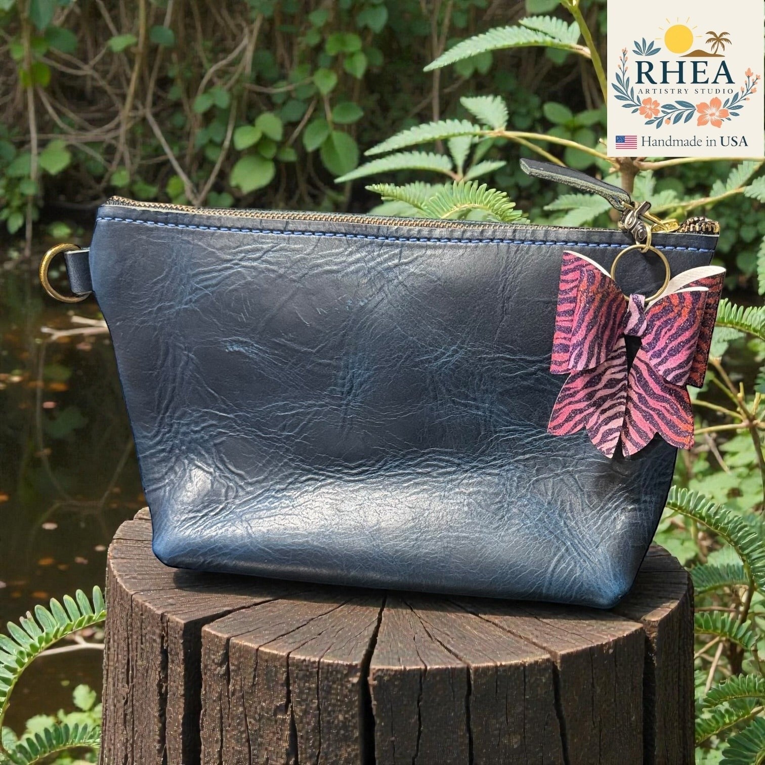 Blue leather pouch with a butterfly purse charm on a wooden stump with green foliage in the background, featuring the RHEA brand.