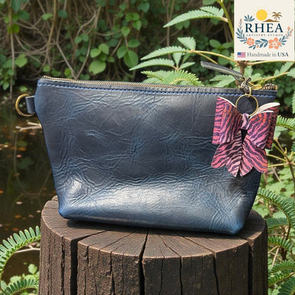 Blue leather pouch with a butterfly purse charm on a wooden stump with green foliage in the background, featuring the RHEA brand.