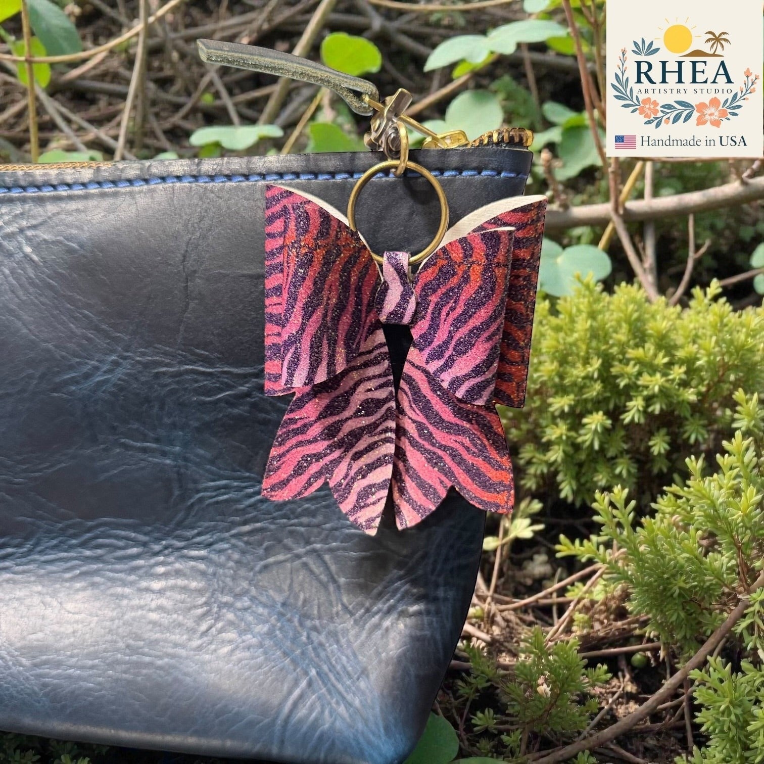 Butterfly-shaped bag charm with pink and purple pattern attached to a black leather bag outdoors.