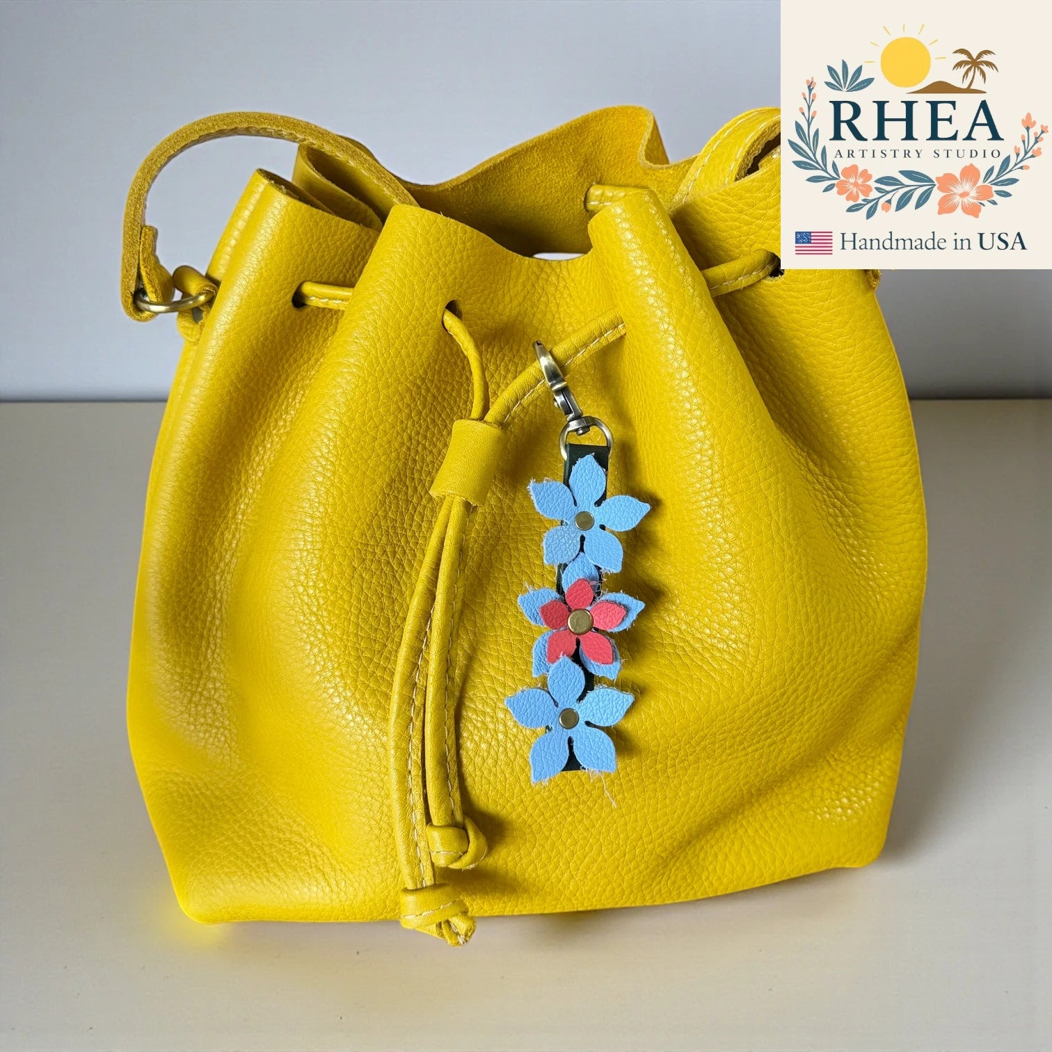Yellow drawstring bag with floral charm on a light gray background, featuring the Rhea Artistry Studio logo.