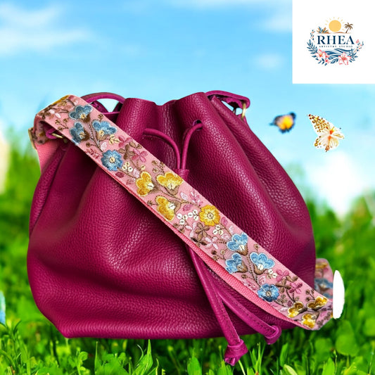 A soft pink embroidered bag strap with whimsical florals in blue, yellow, and brown tones, featuring butterfly-like blooms at garden
