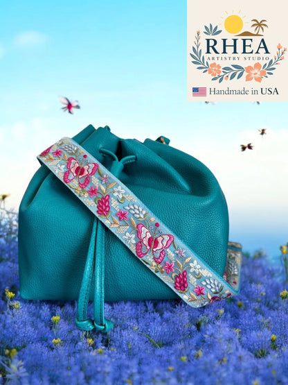 Teal handbag with embroidered purse strap on a blue floral background, featuring Rhea Artistry Studio logo.