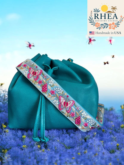 Teal handbag with floral purse strap on a blue background, featuring the RHEA brand.