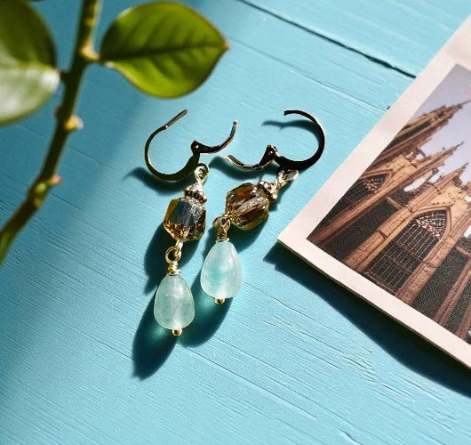 Gold earrings with green teardrop-shaped stones on a blue surface with a blurred architectural background.