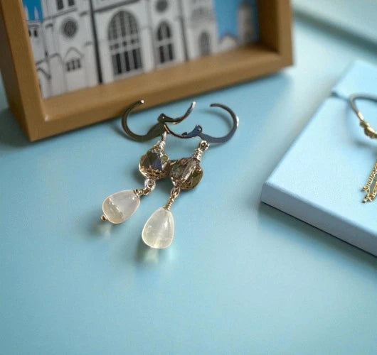 Gold earrings with teardrop-shaped stones on a light blue surface, with a framed picture in the background.
