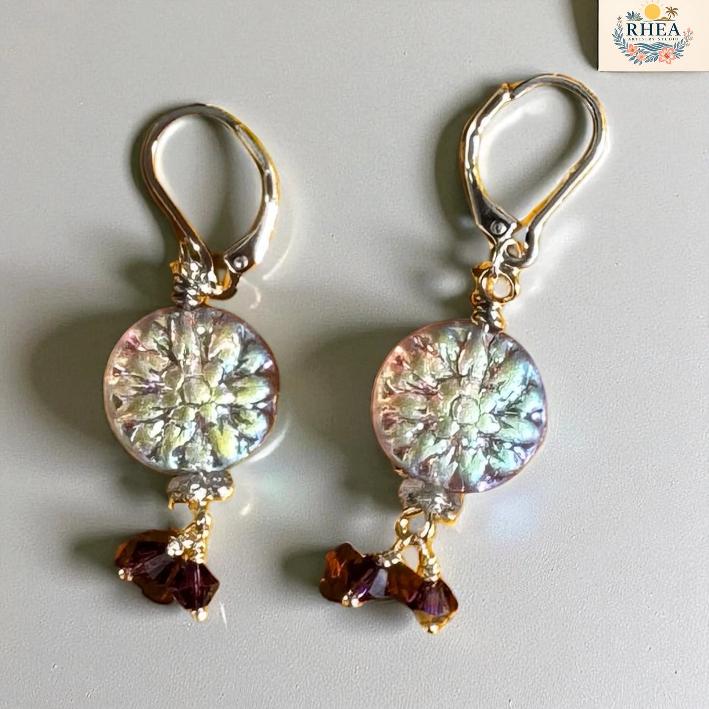 Pair of earrings with floral design and red beads on a light gray background, branded 'RHEA'.