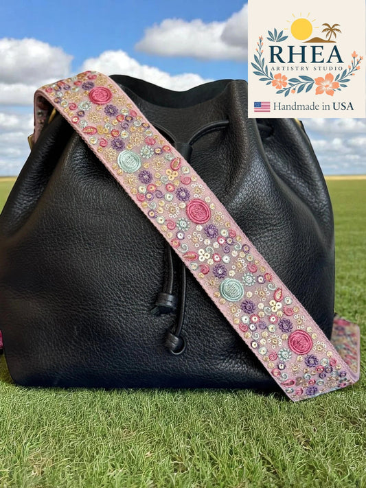 Black leather bag with a floral purse strap on grass with 'RHEA' branding.