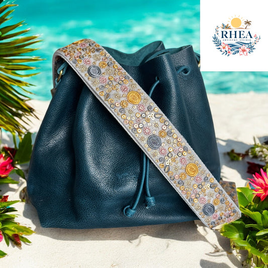 A 2-inch wide strap with gold and silver coin embroidery, sequin florals, and celestial accents, styled with a dark teal leather bag on the beach.