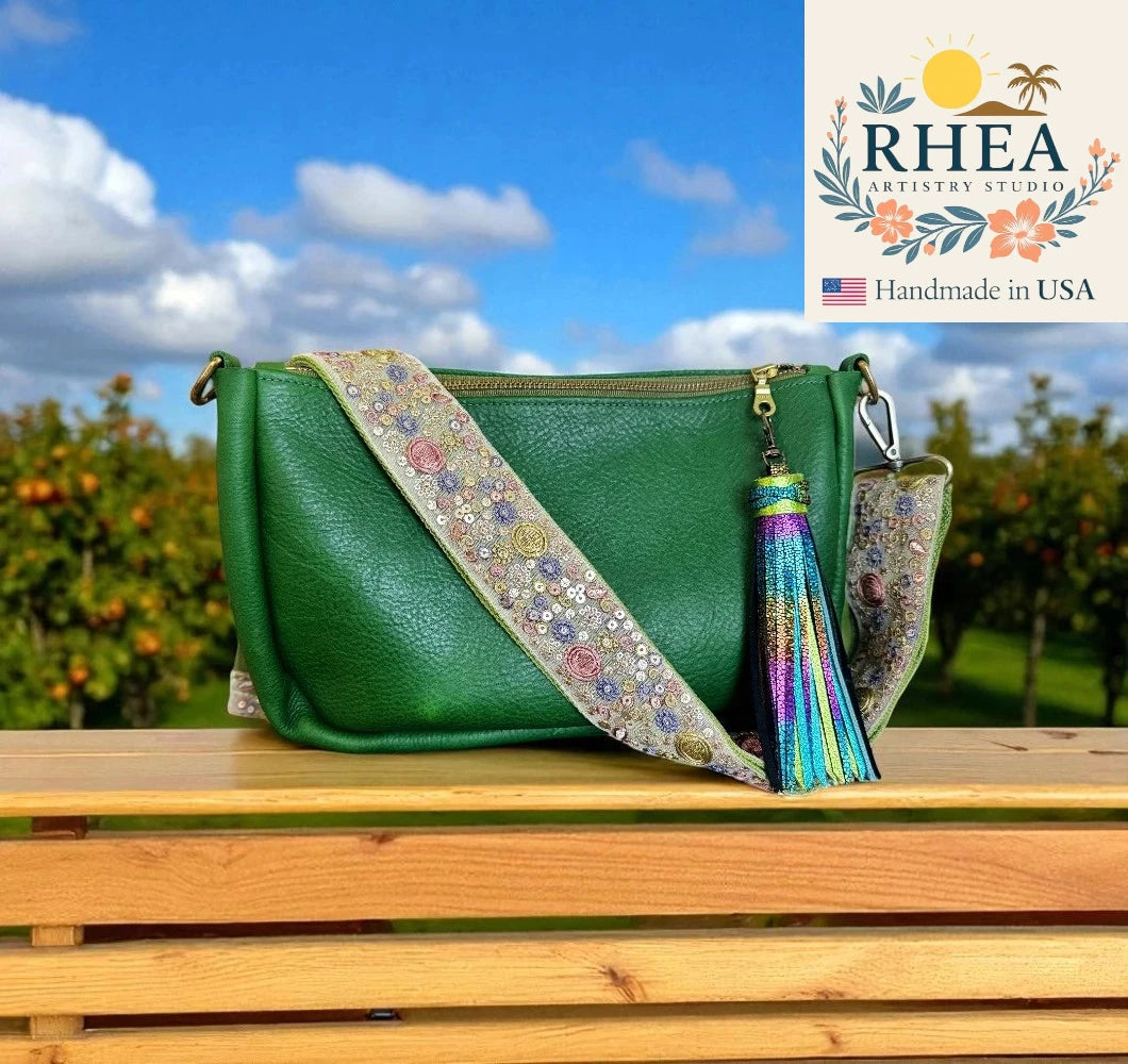 Green handbag with floral purse strap and tassel on a wooden surface with a scenic background, featuring the Rhea Artistry Studio logo.