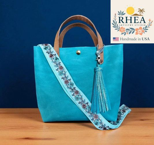 Turquoise handbag with floral bag strap and tassel on a wooden surface, against a blue background with RHEA Artisanry Studio logo.