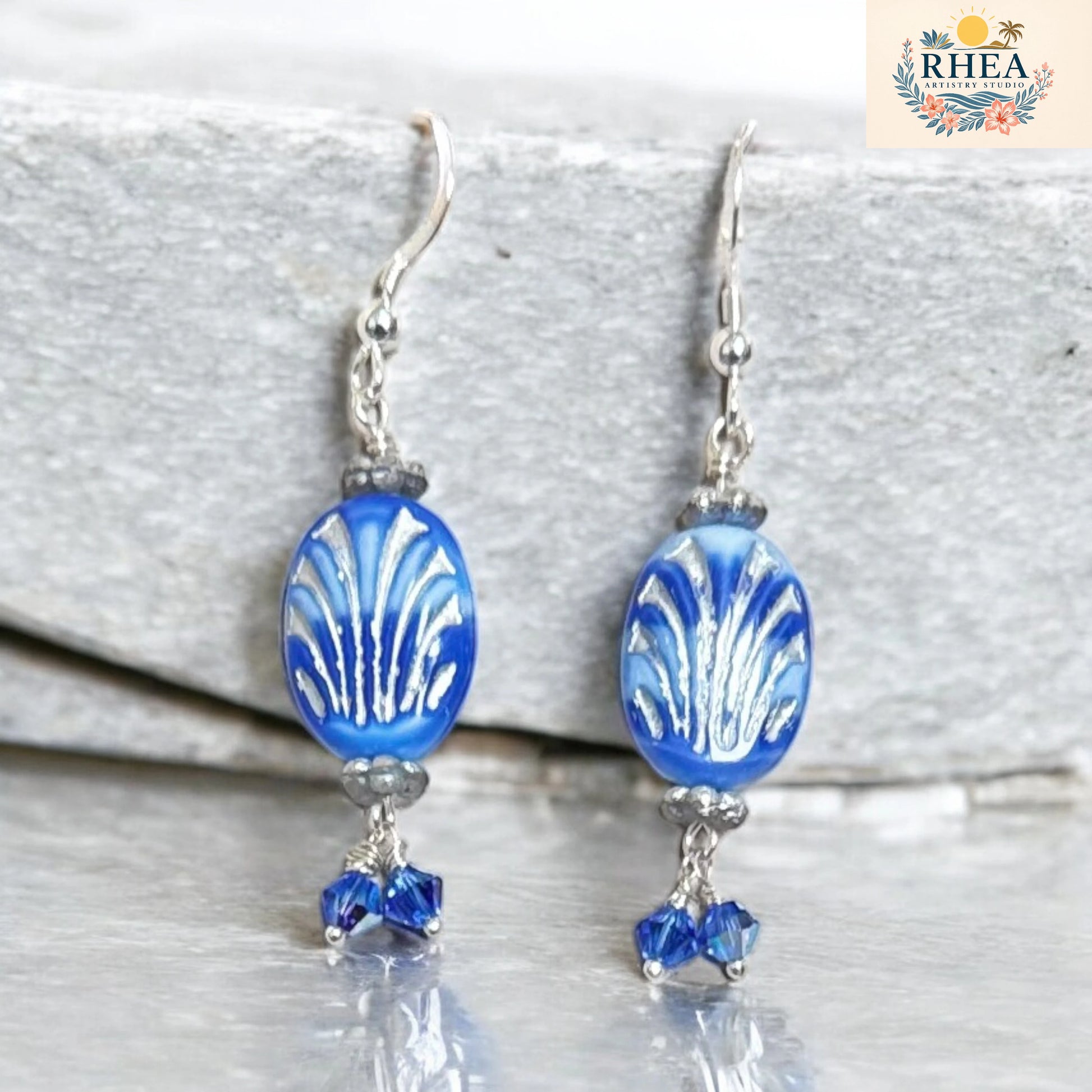 Blue glass bead earrings with silver hooks on a white background, featuring the RHEEA brand logo.
