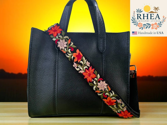 Black handbag with a colorful floral purse strap against a sunset background, featuring the RHEA Artists Studio logo.