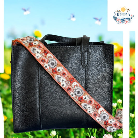 A structured black leather bag paired with a handcrafted strap in coral orange, richly embroidered with detailed floral motifs in black, ivory, and muted red. Surrounded by colorful wildflowers and a bright blue sky, the scene evokes classic botanical illustration with a modern twist.


