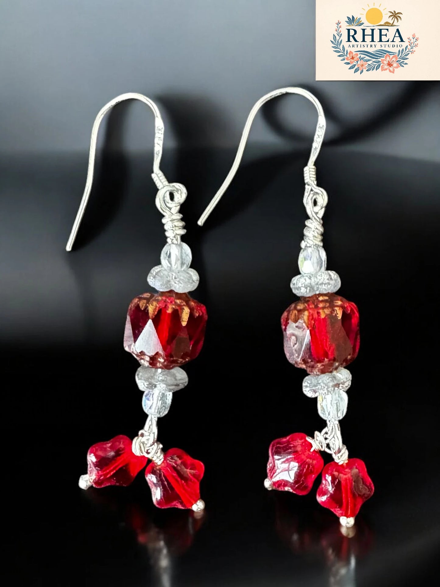 Red and silver beaded earrings on a dark background with RHEA logo.