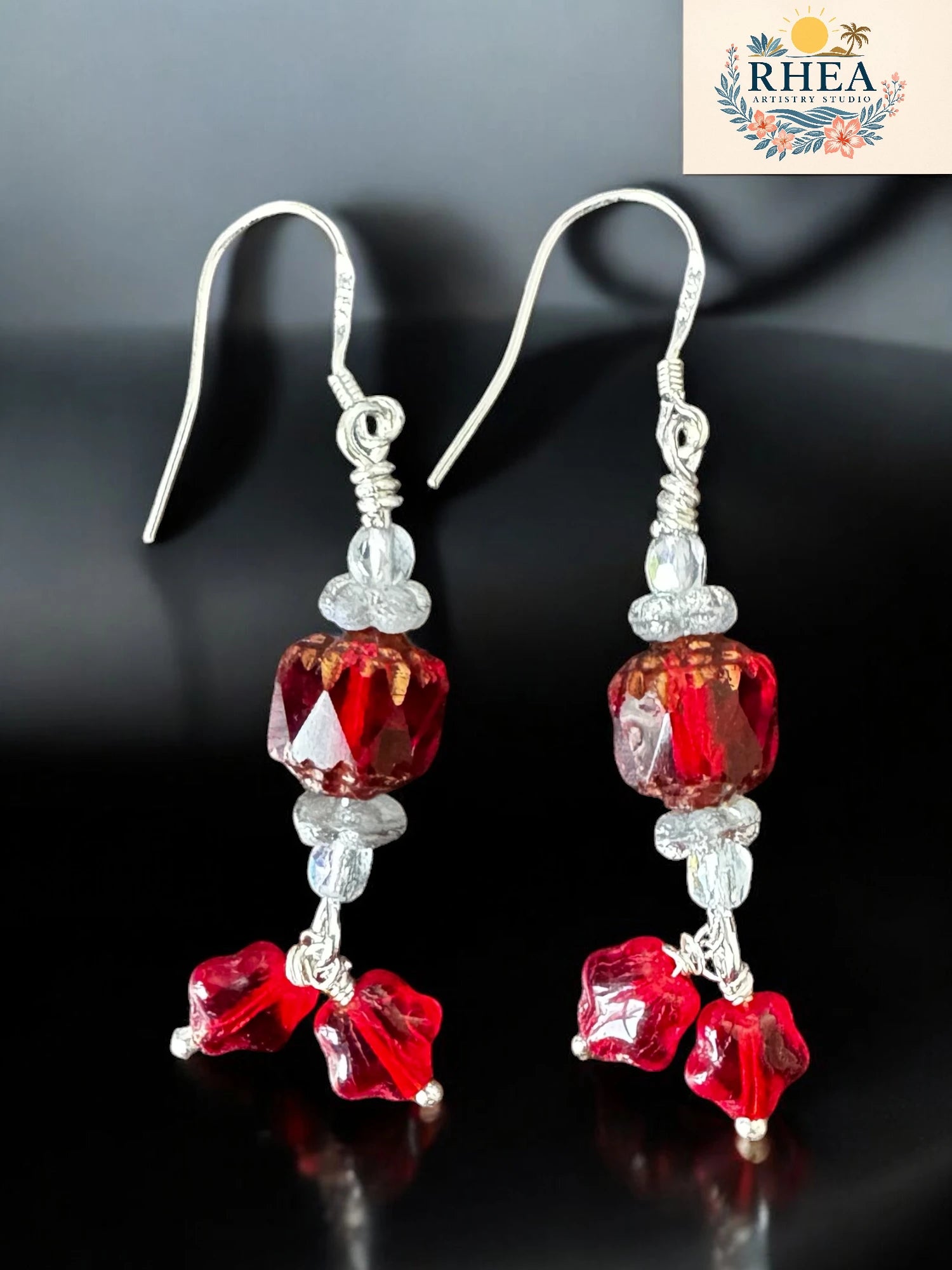 Red and silver beaded earrings on a dark background with RHEA logo.