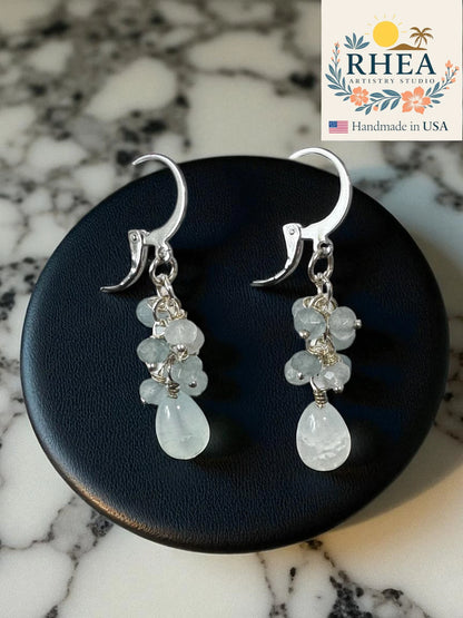 pale blue earrings for women with teardrop-shaped gemstones on a dark surface, branded 'RHEA'.
