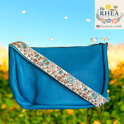 Blue handbag with floral purse strap against a blurred natural background, featuring the RHEA brand.