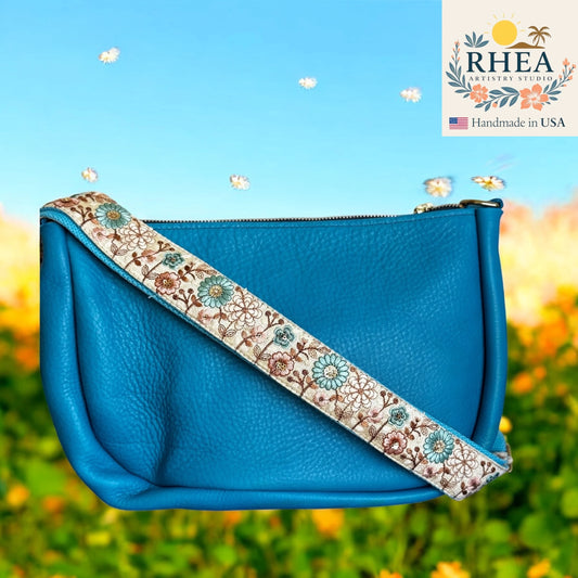 Blue handbag with floral purse strap against a blurred natural background, featuring the RHEA brand.