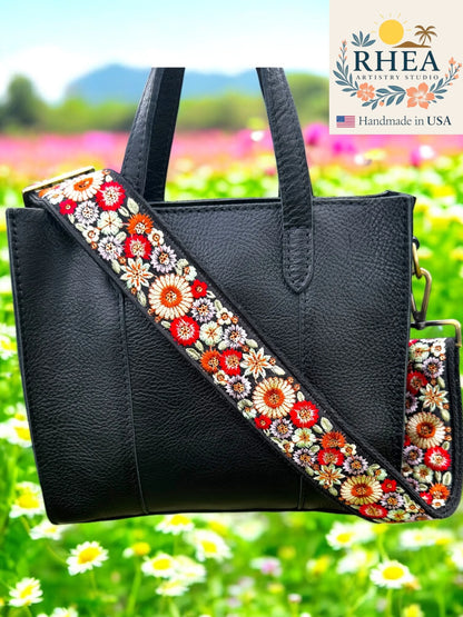 Black handbag with a floral purse strap against a colorful outdoor background, featuring the RHEA brand.
