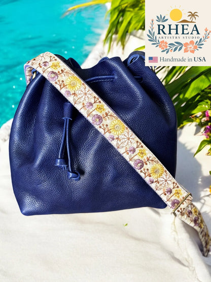 Blue leather drawstring bag with a floral purse strap by RHEA, handmade in USA, on a beach background.