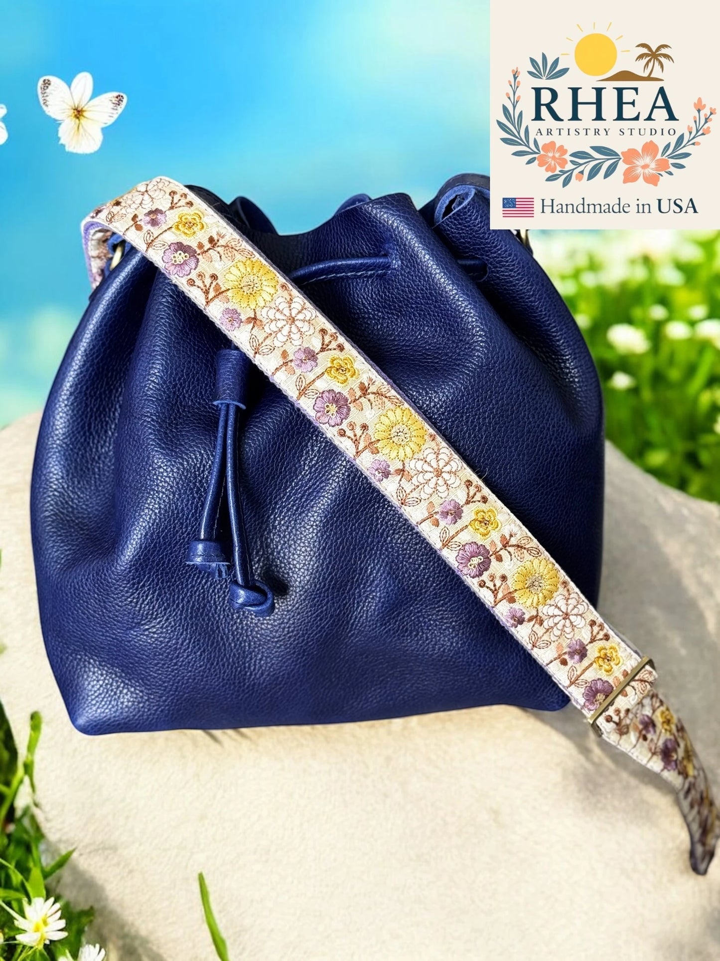 Blue leather drawstring bag with a floral purse strap on a natural background, featuring the RHEA brand.