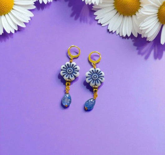 Pair of floral earrings with blue teardrop charms on a purple background with daisies.