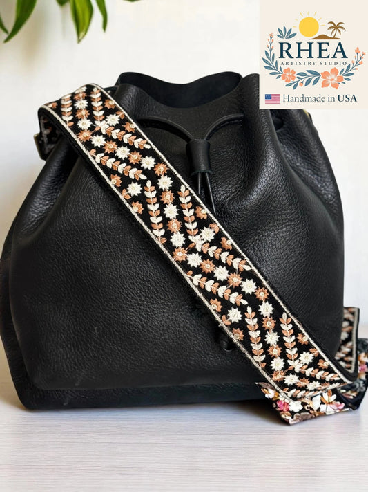 Black leather handbag with a floral purse strap on a light background, featuring the RHEA brand.