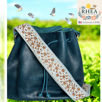 Blue leather bag with an embellished spurse strap against a green outdoor background, featuring the RHEA brand.