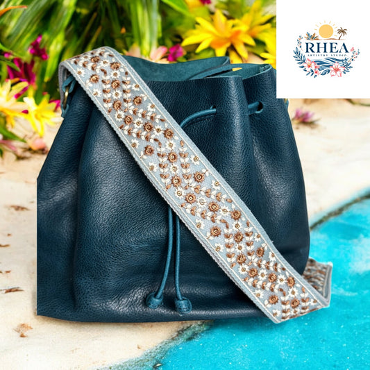 2-inch wide artisan strap featuring brown and bronze embroidered florals on a dusty blue base, styled with a dark navy leather bucket bag near tropical foliage