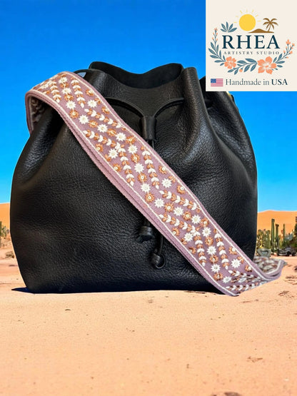 Black leather bag with a decorative purse strap against a desert background, featuring the RHEA brand.